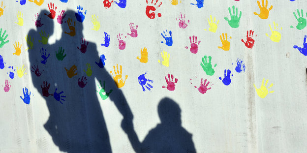 Ein Mann mit einem Kind auf dem Arm und einem an der Hand wirft einen Schatten auf eine mit bunten Handabdrücken bemalte graue Wand.
