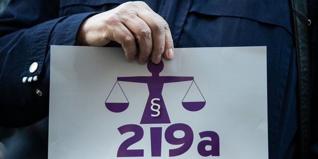 Eine Hand hält ein Schild, auf dem in lila „219a“ steht und eine Waage abgebildet ist