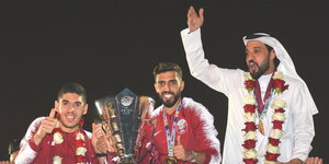 zwei Fußballer halten einen Pokal, daneben steht ein Mann mit erhobenem Arm