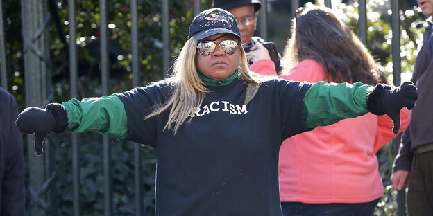 Eine schwarze Demonstrantin trägt ein Shirt mit der Aufschrift „Rassismus“ und zeigt die Daumen nach unten
