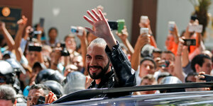 Ein Mann mit schwarzem Bart, gegelten Haaren und schwarzer Lederjacke winkt aus einem Auto.
