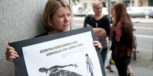 Demonstrantin hält Schild mit Aufschrift: "Genitalverstümmelung verfolgt ein Leben lang"