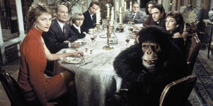 Bei dem Foto aus einem Film sitzen zahlreiche schick gekleidete Menschen am Essenstisch, gemeinsam mit einem Gorilla