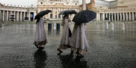 Drei Nonnen laufen bei Regen mit Regenschirmen über einen Platz