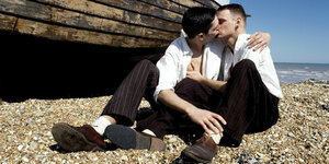 Zwei Männer in weißen Hemden sitzen am Strand und küssen sich.