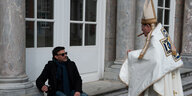 Ein Priester mit Zigarre kommt auf einen sitzenden Mann zu