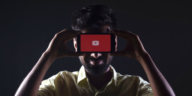 Mann hält sich Smartphone vor die Augen, Display zeigt zum Betrachter - auf ihm ist das Youtube-Logo zu sehen