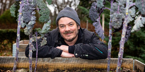 Ein Mann mit Mütze lehnt an einem Hochbeet mit Gemüse und lächelt