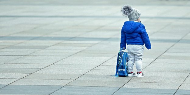 Ein einzelnes Kind mit einem Rucksack in der Hand steht auf einem Platz