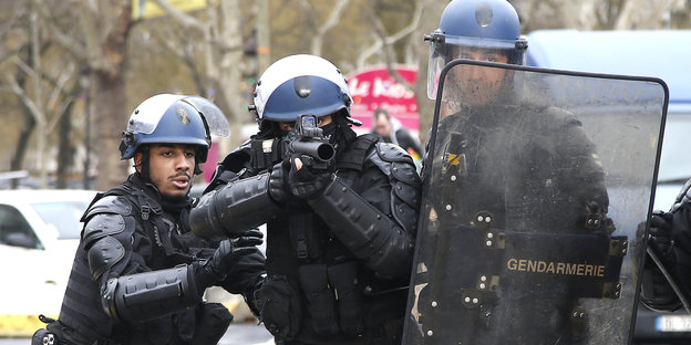 Drei Polizisten, einer von ihnen zielt mit einem Tränengasgewehr in Richtung Fotograf