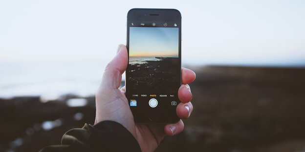 Eine Hand hält ein Smartphone und macht ein Foto vom Meer, das auch im Hintergrund zu sehen it
