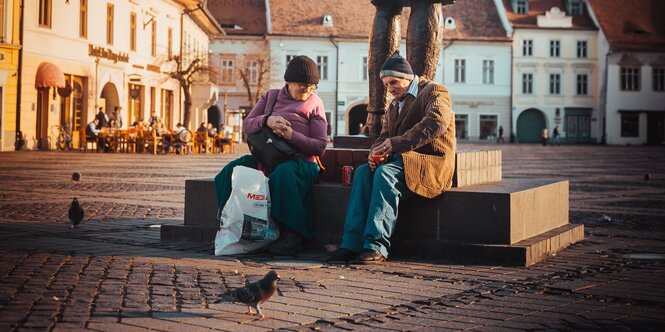 Zwei ältere Menschen, ein Mann und eine Frau, sitzen auf einem Platz und trinken Cola aus Dosen