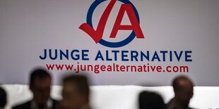 Menschen stehen vor einem wandfüllenden Logo der "Jungen Alternative".