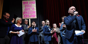 Menschen auf einer Theaterbühne halten ein Schild mit der Aufschrift: "Wir sind viele - jedeR einzelne von uns"