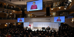 Der Saal der Sicherheitskonferenz mit Angela Merkel auf der Bühne auf drei großen Bildschirmen