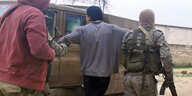 Ein Mann steht zwischen zwei bewaffneten Männern in Uniformen und wird von ihnen festgehalten