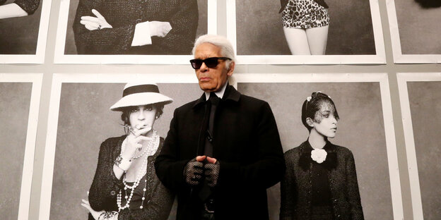 Karl Lagerfeld steht vor Porträtaufnahmen, er trägt eine schwarze Brille und schwarze Handschuhe
