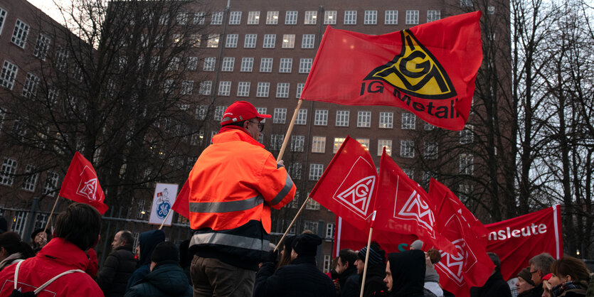 Viele Menschen stehen vor einem vielstöckigen Gebäude und protestieren mit Fahnen der Gewerkschaft IG Metall