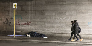 Zwei Menschen laufen unter einer Brücke, vor ihnen liegt ein Obdachloser auf dem Boden