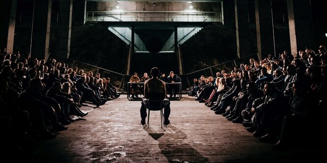 Filmszene: Ein Mann sitzt in einem Raum in der Mitte. Sehr viel leute sitzen um ihn herum und schauen ihn an