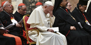Papst Franziskus sitzt auf einem Stuhl und hält sich die linke Hand vors Gesicht