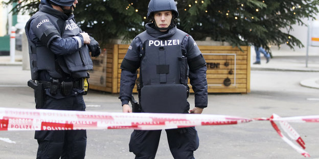 Zwei Polizisten stehen in Schutzkleidung hinter einem Flatterband auf einem Weihnachtsmarkt
