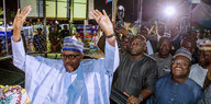 Buhari hebt jubelnd die Hände in die Höhe, im Hintergrund stehen mehrere Männer, darunter ein Fotograf und Polizisten.