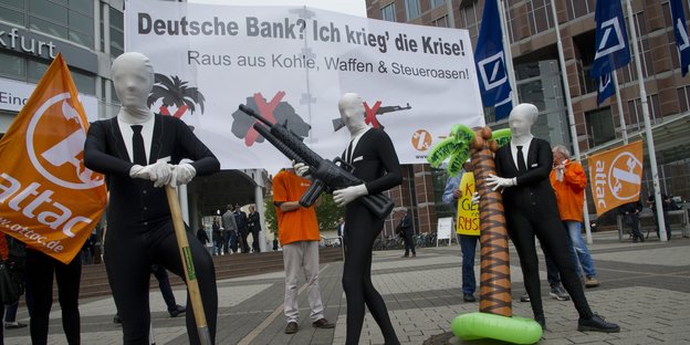 Menschen in Anzügen und mit weißen Masken protestieren mit Schildern vor der Deutschen Bank in Frankfurt.