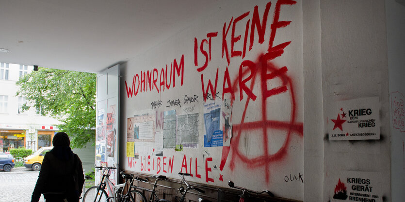 Frau geht an Graffiti gegen Immobilienspekulation an Häuserwand vorbei.