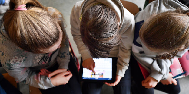 Drei Schulkinder, von oben fotografiert, sitzen nebeneinander. Das in der Mitte hält ein Tablet in der Hand.