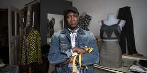 Ein dunkelhäutiger Mann in Jeansjacke mit Basecap auf dem Kopf und Metermaß in der Hand steht zischen Nähatelierpuppen, auf denen halb fertige Kleider hängen