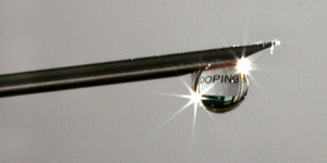 an der Spitze einer Kanüle hängt ein Tropfen auf dem sich das Wort „Doping“ spiegelt