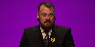 Mark Meechan trägt einen Ukip-Button und steht vor einer lilafarbenen Wand