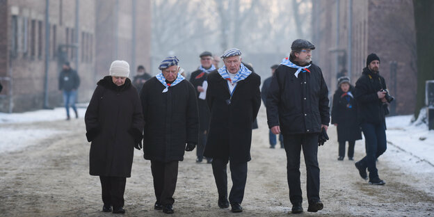 Alte Männer und Frauen mit Häftlingsmützen gehen über das Auschwitz-Gelände.