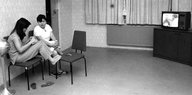 Zwei Frauen sitzen auf Stühlen in einem kargen Aufenthaltsraum, in einer Ecke läuft ein Fernseher