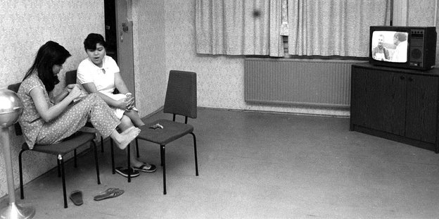 Zwei Frauen sitzen auf Stühlen in einem kargen Aufenthaltsraum, in einer Ecke läuft ein Fernseher