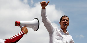 Guaidó in Sprecherpose gegen den blauen Himmel. In das Foto ragt ein Arm, die Hand hält ein Megaphon