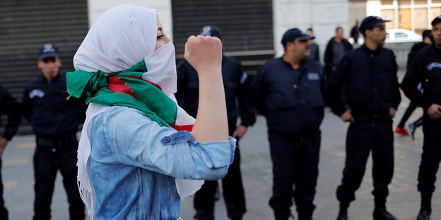 Eine Frau mit Kopftuch und algerischer Flagge um den Hals ballt die Hand zur Faust. Im Hintergrund stehen uniformierte Männer in einer Reihe.