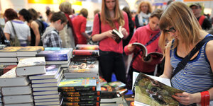 Jugendliche Mädchen stehen auf der Buchmesse vor Bücherstapeln und lesen jeweils in einem Buch