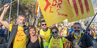 zwei Frauen und zwei Männer stehen Arm in Arm nebeneinander, im Hintergrund eine Demonstrationszug