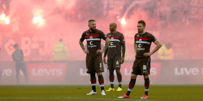 Drei Spieler des FC St. Pauli stehen auf dem Spielfeld des Millerntor-Stadions, im Hintergrund qualmen Bengalos.