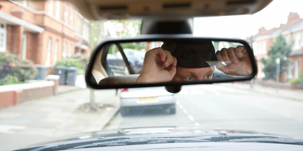 Ein Mann mit Mütze blickt vom Inneren eines Autos in einen Rückspiegel.