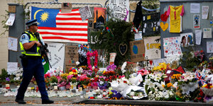 Ein Polizist mit Gewehr im Anschlag läuft an einem Zaun vorbei, vor dem Blumen und Kränze niedergelegt sind und an dem Fotos und Bilder hängen
