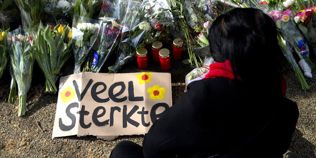 Eine Frau legt Blumen nieder. Auf dem Boden liegt ein Schild, auf dem auf Niederländisch "Viel Kraft" steht.