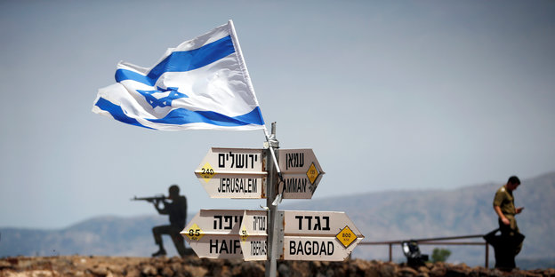 Ein Wegweiser mit Pfeilen in verschiedene Richtungen, darüber weht die israelische Flagge. Im Hintergrund sind Soldaten zu sehen