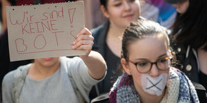 Eine Frau mit zugeklebtem Mund und eine, die en Plakat hochhält, laufen auf einer Demonstration