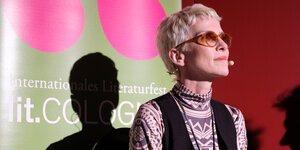 Eine blonde Frau mit Sonnenbrille vor einer Stellwand.
