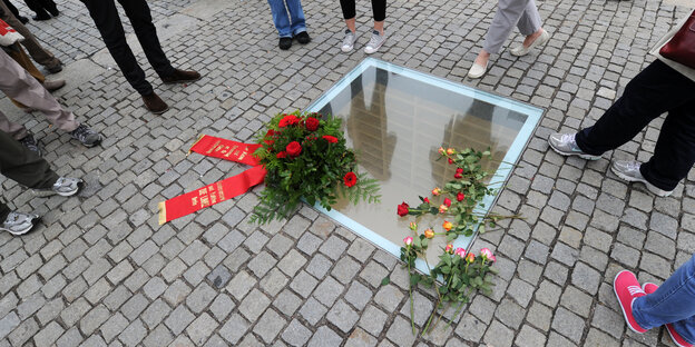 Blumenkränze liegen auf einer eingelassenen Scheibe auf einem gepflasterten Platz