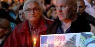 Zwei Männer halten eine Kerze und ein Plakat bei einer Gedenkveranstaltung zu den Nato-Angriffen.