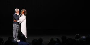 Apple-Chef Tim Cook und Moderatorin Oprah Winfrey umarmen sich vor einem schwarzen Hintergrund
